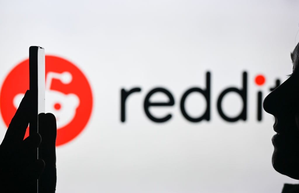 An image of a woman holding a cell phone in front of the Reddit logo displayed on a computer screen. Image used for SmartBrief article with headline: Why it’s time to add Reddit marketing to your mix